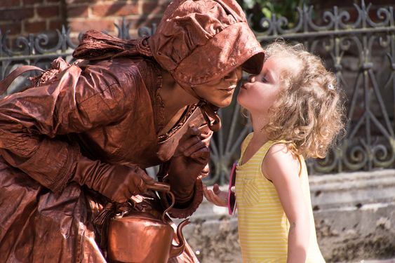 Miss Copper Kettle - a Victorian, realistic-looking living statue that leans in while a blond girl in the yellow, summery t-shirt kisses her on the cheek. Photo is taken at the Statue-En Marche in Belgium.