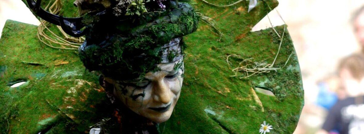 Nature Goddess, adorned with a forest crown and moss collar, stands at an eco and environmental event. A child shows a delicate, white flower to the Nature character adding to the enchanting atmosphere. The image captures the joy and wonder of this unique performance, celebrating nature and inspiring a sense of awe in all who witness it.