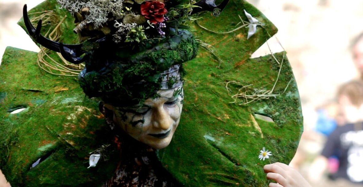Nature Goddess, adorned with a forest crown and moss collar, stands at an eco and environmental event. A child shows a delicate, white flower to the Nature character adding to the enchanting atmosphere. The image captures the joy and wonder of this unique performance, celebrating nature and inspiring a sense of awe in all who witness it.