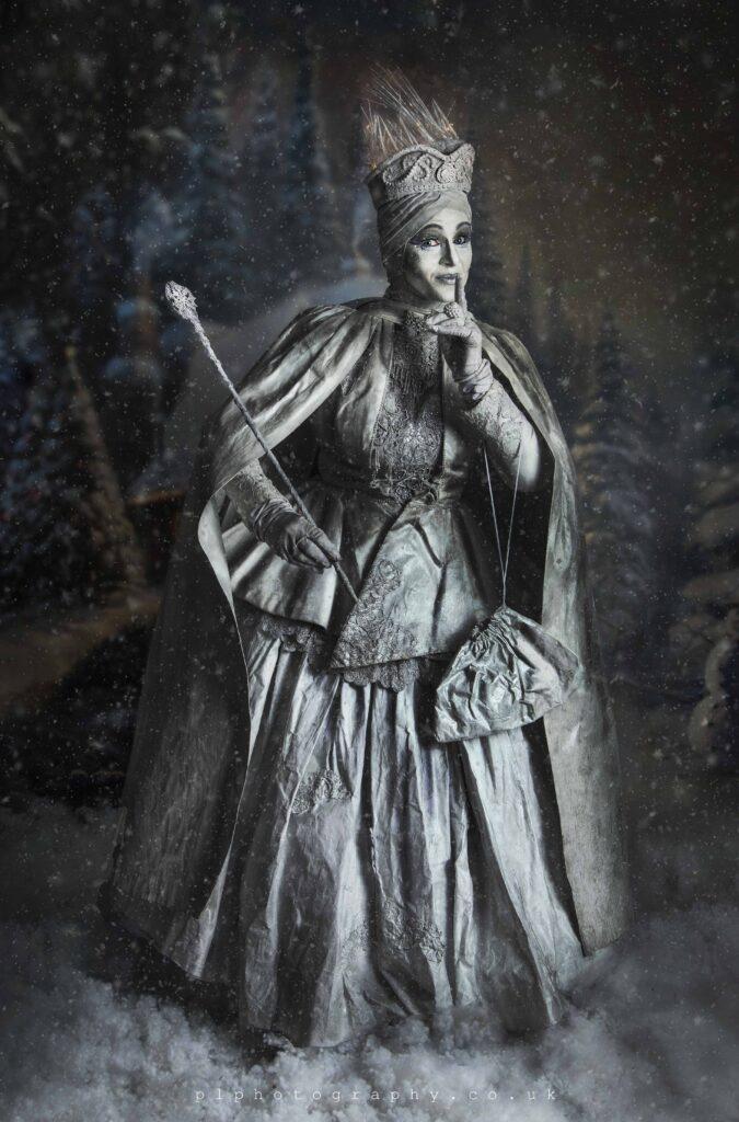 Enchanting Snow Queen amidst snowy scenery in her impressive Victorian costume, adorned with a long dress, cape, and crown. She holds a wand in her hand and places a finger to her lips, gesturing to keep silence. Captured in Dunbar, Scotland by plphotography.co.uk