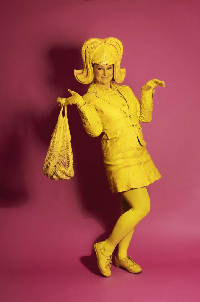Lemon Lush is a whimsical character. She holds a bag with bananas, her expression radiating joy. Adorning her head is a large, yellow wing, complementing her vibrant costume. She wears a playful short dress paired with a jaunty jacket.