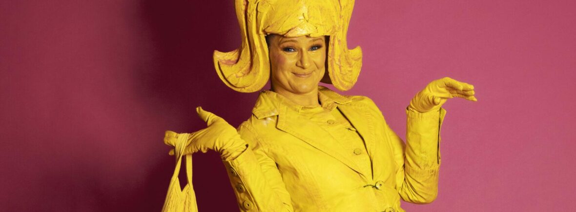 Lemon Lush is a whimsical yellow living statue holds a bag with bananas, her expression radiating joy. Adorning her head is a large, yellow wing, complementing her vibrant costume. She wears a playful short dress paired with a jaunty jacket.