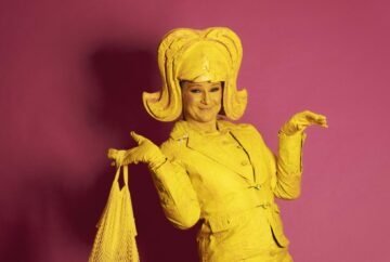 Lemon Lush is a whimsical yellow living statue holds a bag with bananas, her expression radiating joy. Adorning her head is a large, yellow wing, complementing her vibrant costume. She wears a playful short dress paired with a jaunty jacket.
