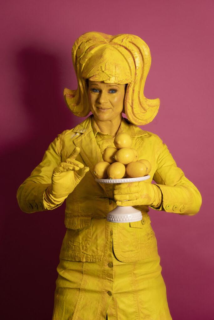 Lemon Lush is a whimsical yellow living statue holds a plate with lemons, her expression radiating joy. Adorning her head is a large, yellow wing, complementing her vibrant costume. She wears a playful short dress paired with a jaunty jacket.