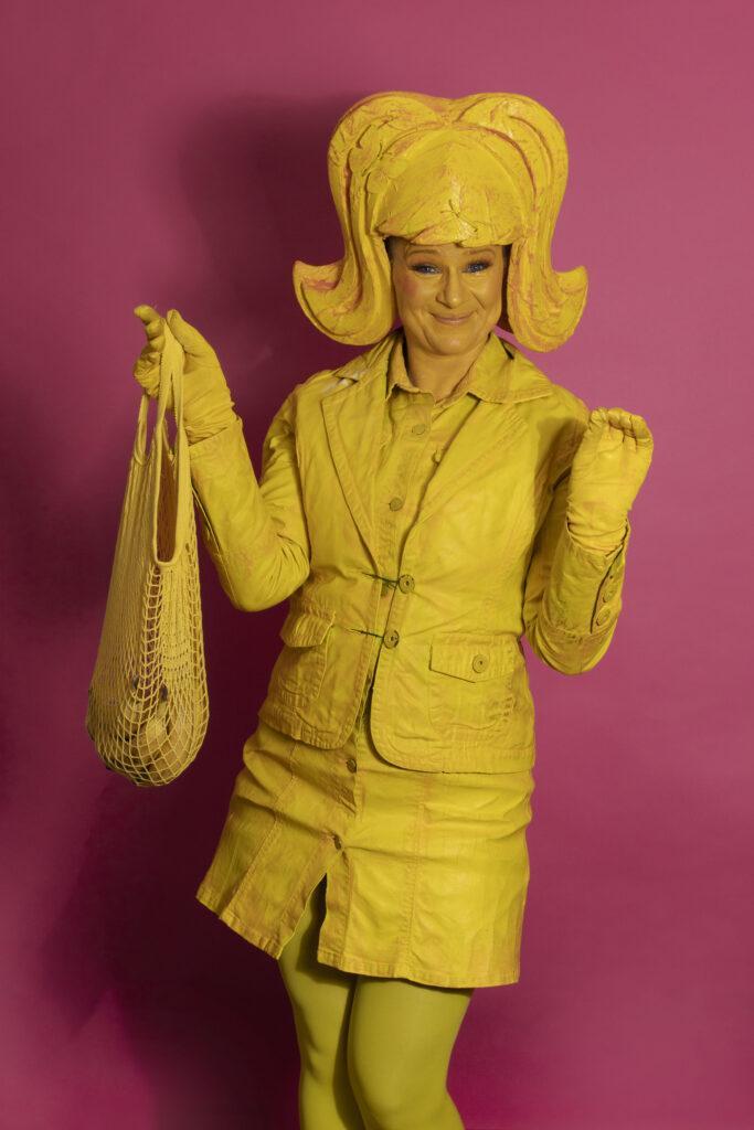 A whimsical yellow living statue holds a bag with bananas, her expression radiating joy. Adorning her head is a large, yellow wing, complementing her vibrant costume. She wears a playful short dress paired with a jaunty jacket.