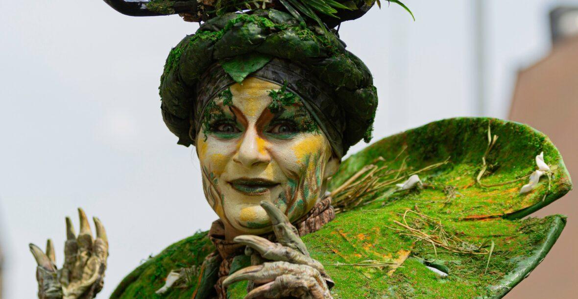 Nature Goddess, adorned with a forest crown and moss collar, stands at an eco and environmental event. She looks strait at you raising her claws as if she wanted to say something. The image captures the joy and wonder of this unique performance at Lommel festival, celebrating nature and inspiring a sense of awe in all who witness it. It is an intriguing roaming act for events.