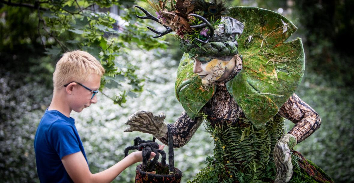 A Nature Goddess at the festival shows a spider to a curious boy, surrounded by greenery and woodland creatures.