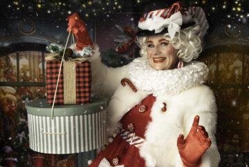 Elegant Christmas elf in a red dress with white satin collar and fluffy white jacket, smiling while presenting Christmas gifts at a festive holiday event. Styled in a vintage aesthetic, the elf has a Marilyn Monroe-inspired hairstyle, adding a touch of classic glamour to the celebration.