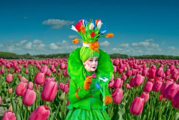 A whimsical character named Miss Tulip stands facing the viewer in a field of blooming pink tulips, smiling playfully against the colorful floral background.